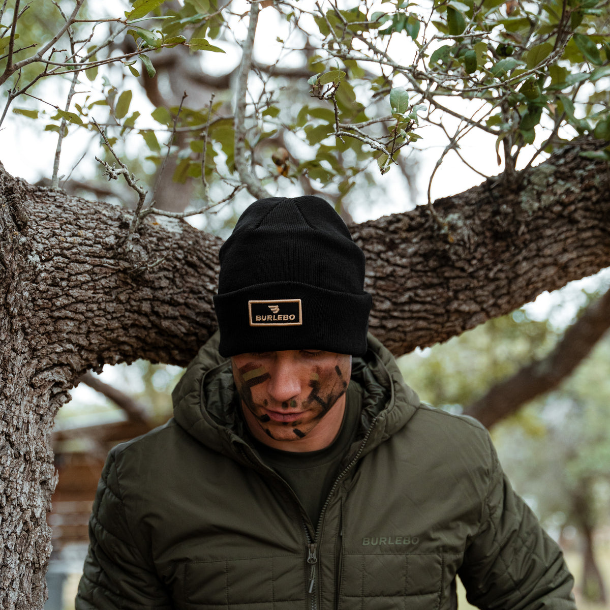 Man with face paint wearing the Black BURLEBO Beanie and an olive jacket under an oak tree