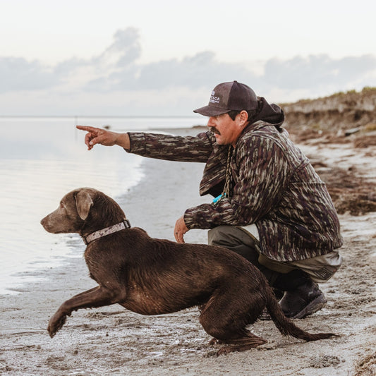 Man in camouflage jacket and cap with a brown dog by a body of water