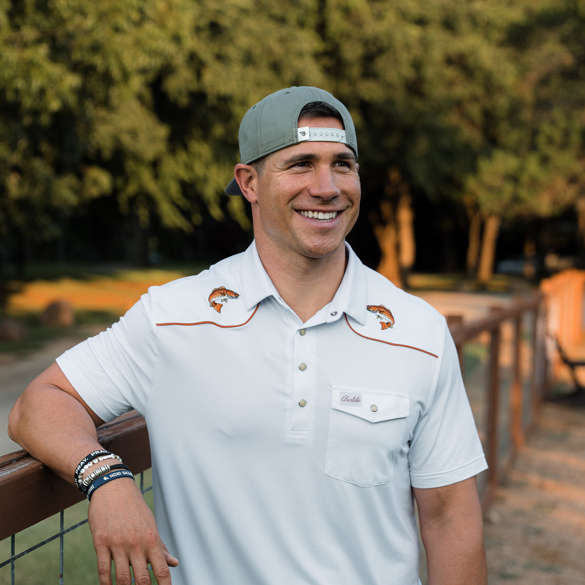 Man wearing a light gray polo shirt with a logo, standing outdoors with trees in the background.