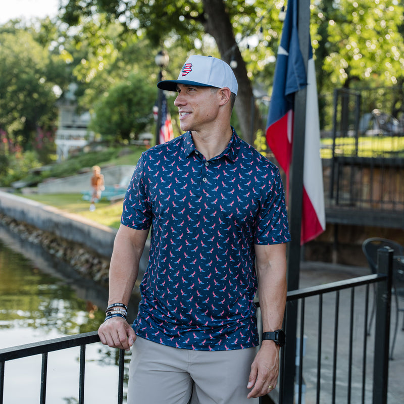 Man wearing a patterned polo shirt and cap standing by a waterfront with flags in the background