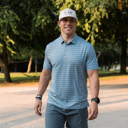 Man wearing a blue checkered shirt and white cap standing outdoors with trees in the background