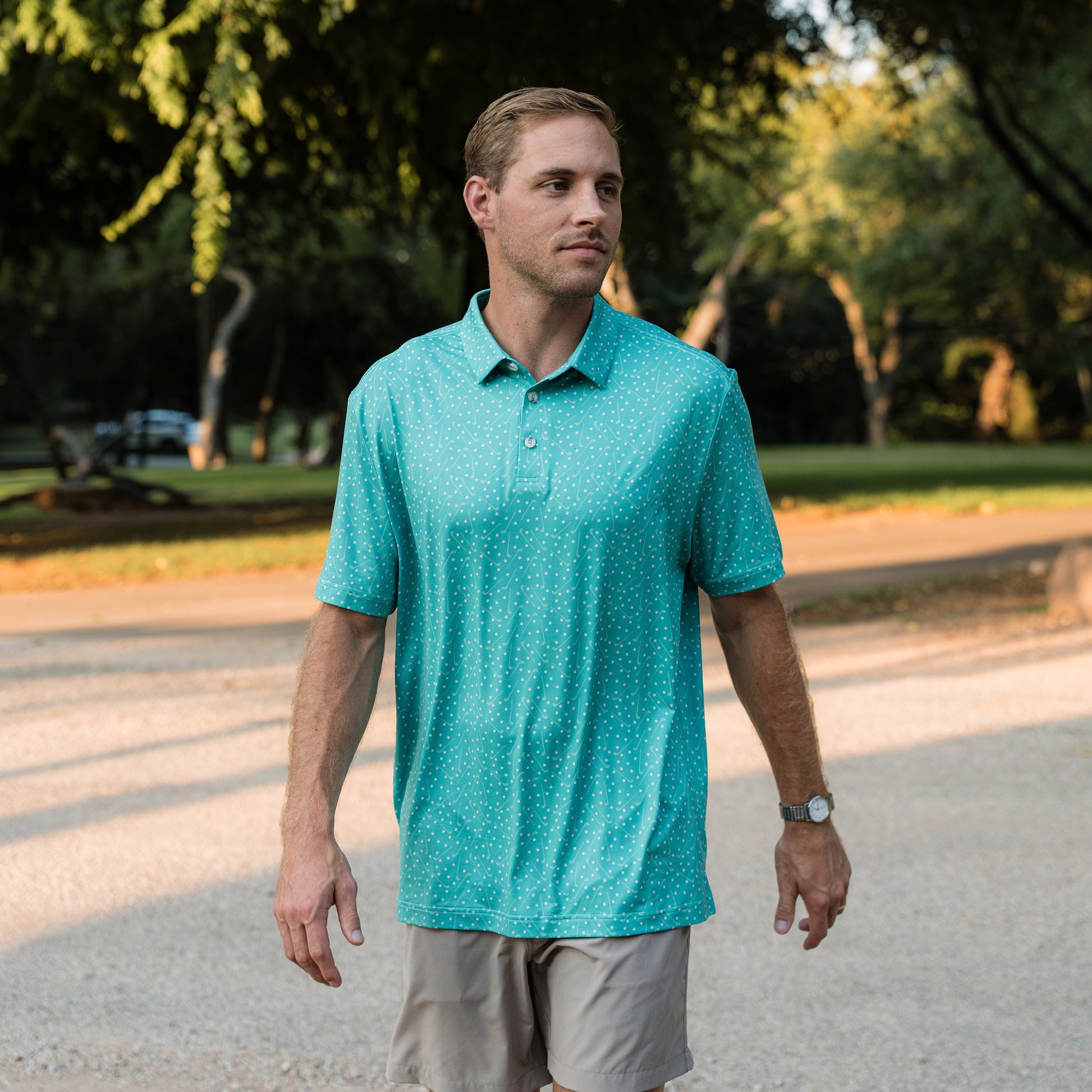 Man wearing a teal polo shirt walking outdoors on a path with trees in the background