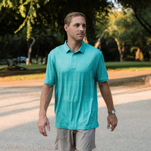 Man wearing a teal polo shirt walking outdoors on a path with trees in the background