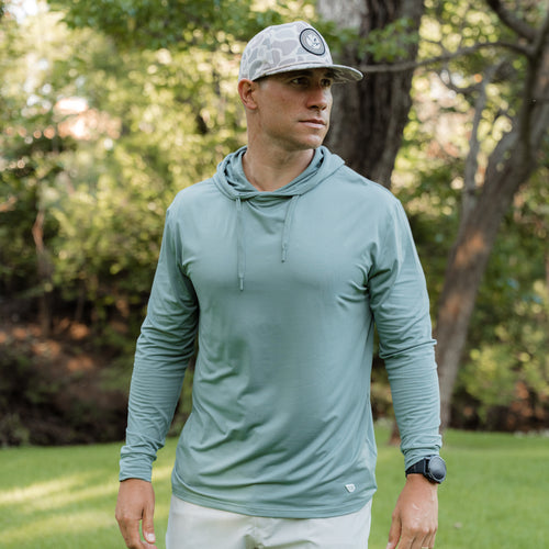 Man wearing a light green long-sleeve hoodie and cap outdoors in a park setting