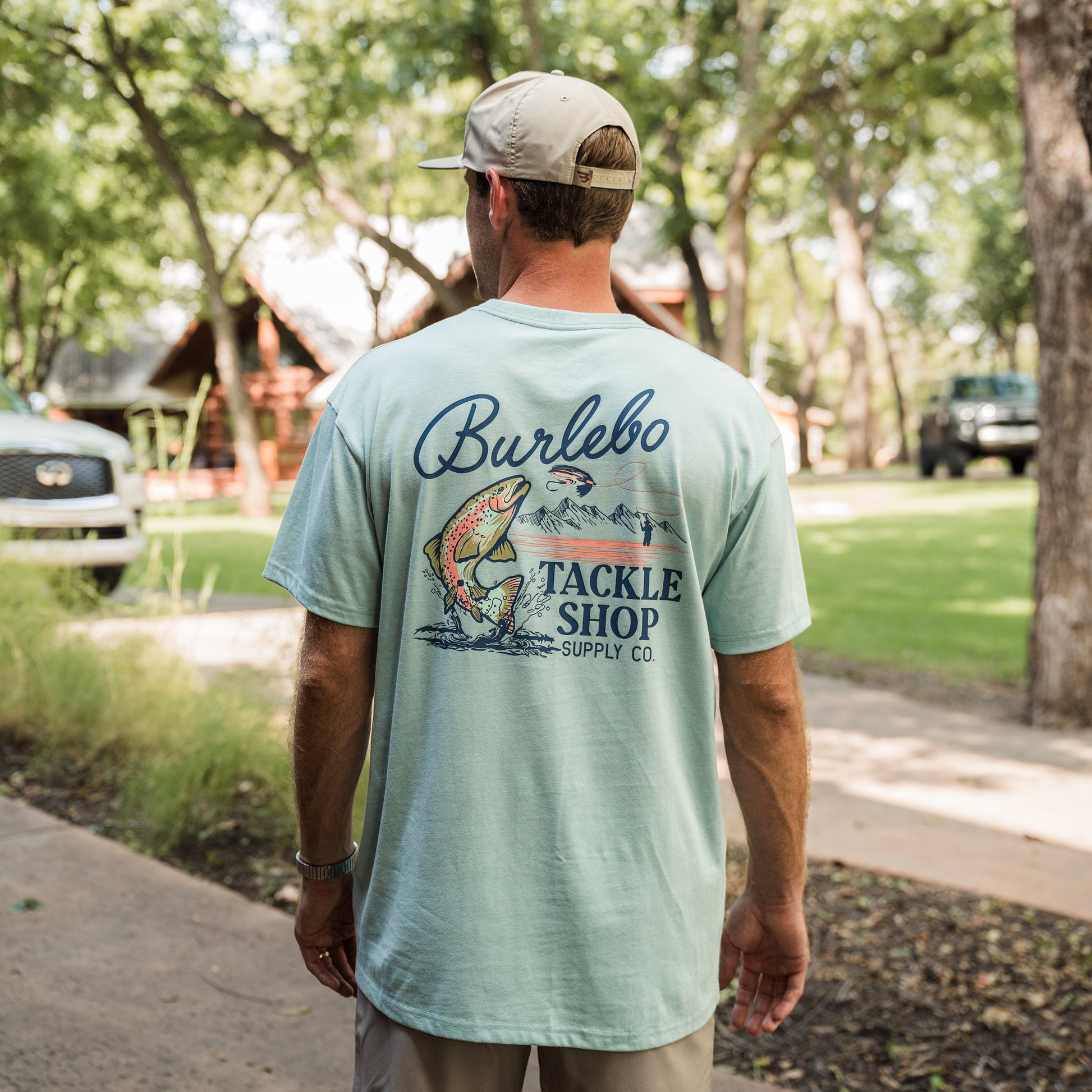 Man wearing a light blue t-shirt with 'Burlebo Tackle Shop' design, walking outdoors.