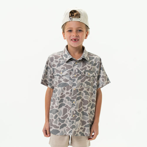 Child wearing a camouflage-patterned shirt and cap on a white background