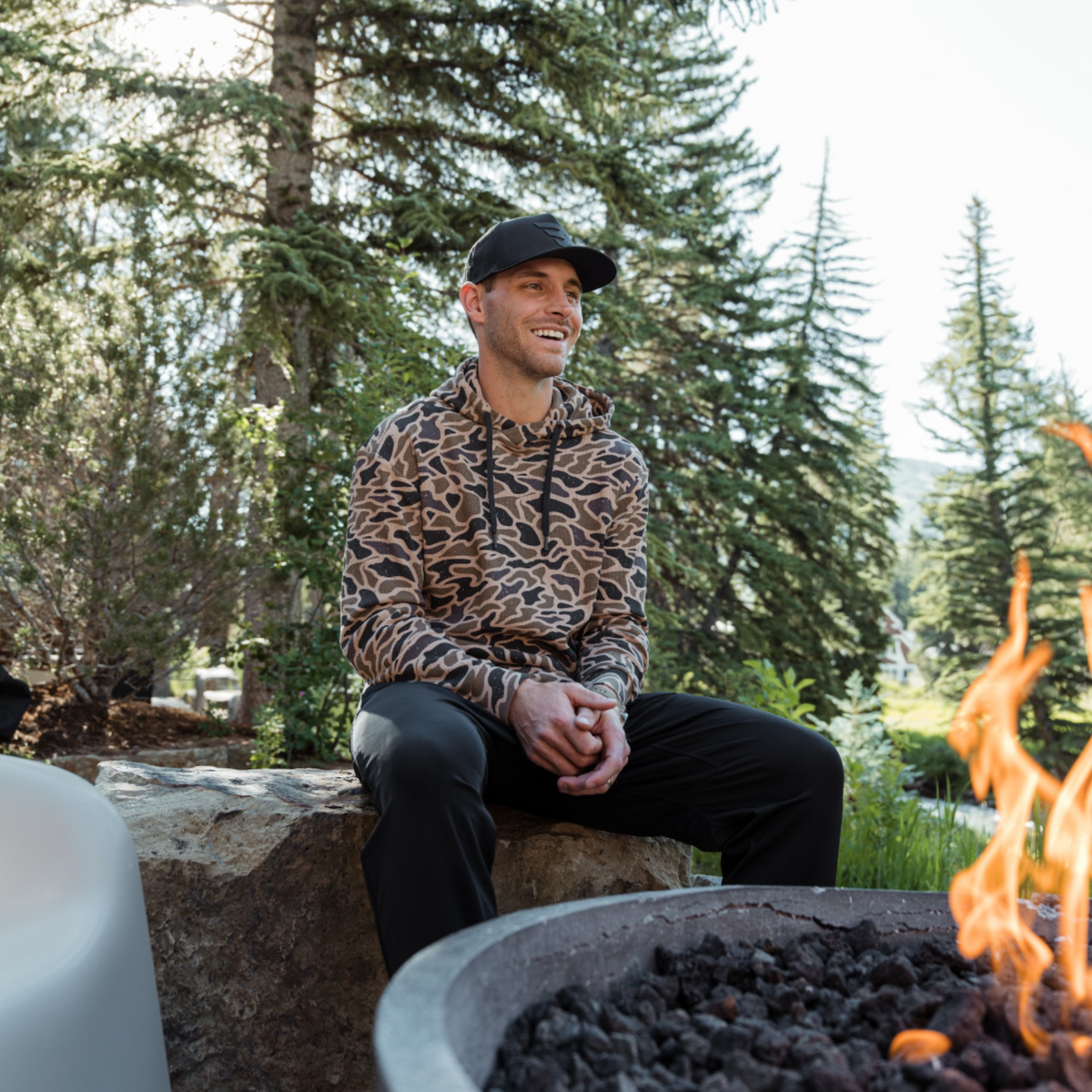 Smiling man seated by an outdoor fire pit among pine trees in the Gauge Camo Thermal Hoodie