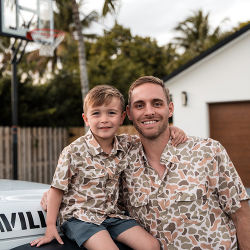 Man and young boy wearing matching patterned shirts outdoors with a basketball hoop in the background.