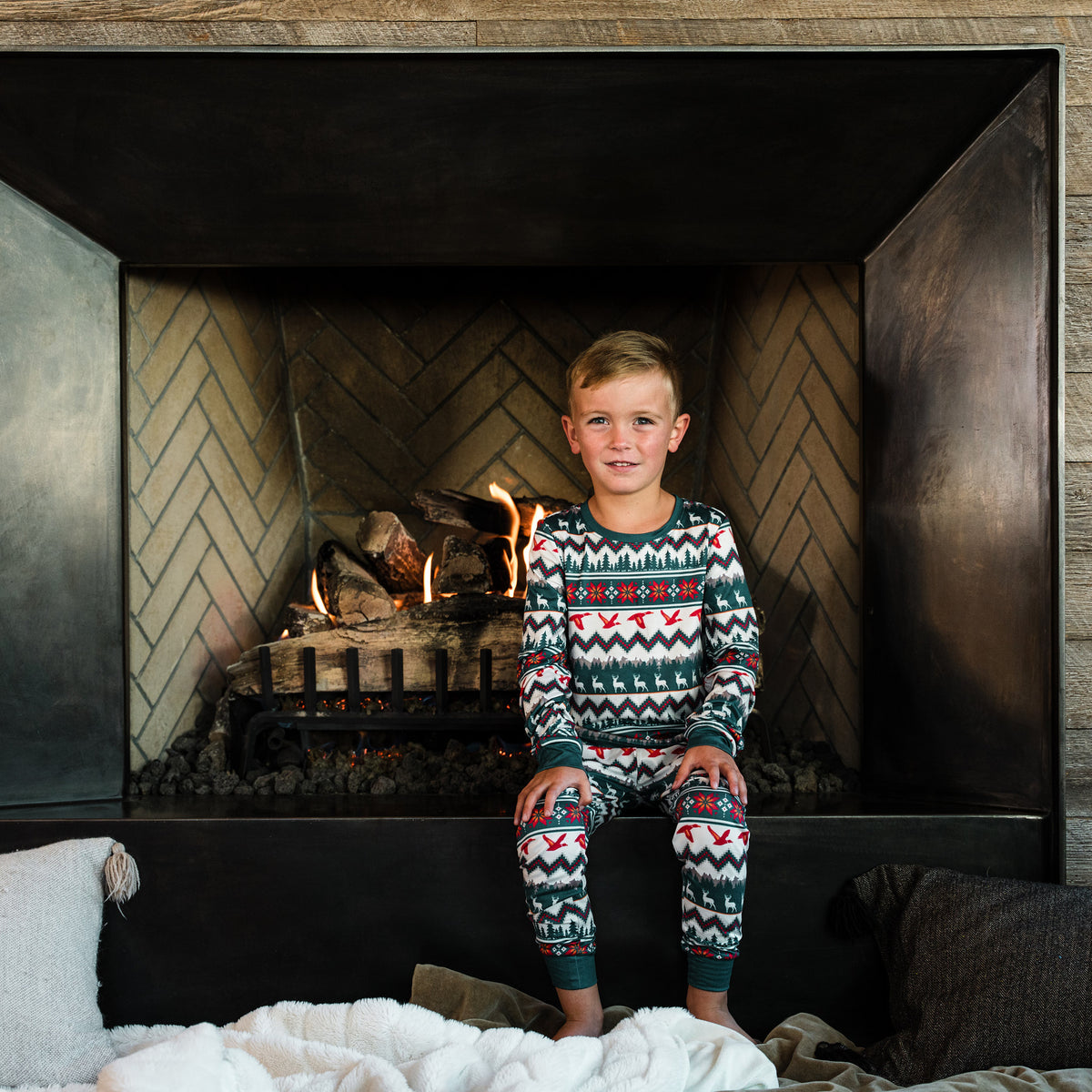 Boy in the BURLEBO Outdoors Christmas Youth Pajama Set sitting in front of a lit fireplace