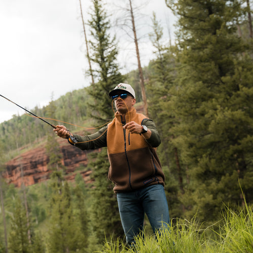 Man in the Multi Brown Montana Vest and a white cap casting a fly rod on a grassy hillside by pine trees