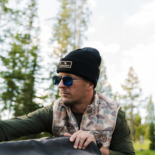 Man in sunglasses and a camo vest wearing the Black BURLEBO Beanie outdoors among pine trees