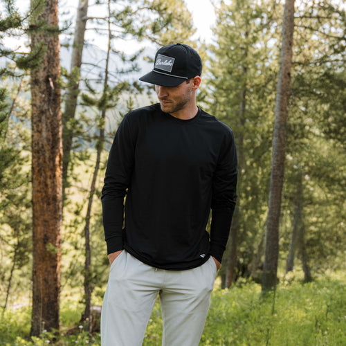 Man in a black cap wearing the Black Long Sleeve Go To Tee and light gray pants in a pine forest