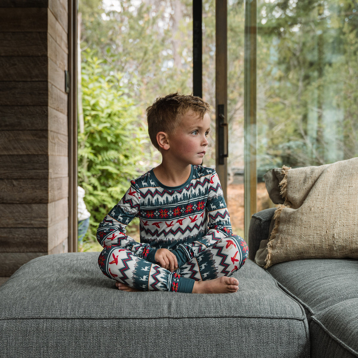 Boy in the BURLEBO Outdoors Christmas Youth Pajama Set sitting cross-legged on a gray couch by a window