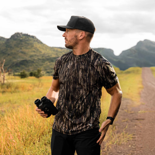 Man in camouflage shirt and cap holding binoculars in a natural setting with mountains.